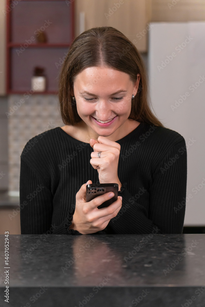 Overwhelmed with joy, a young woman bites her fingers while reading a message on her phone in a modern kitchen, expressing genuine happiness and excitement. Vertical photo