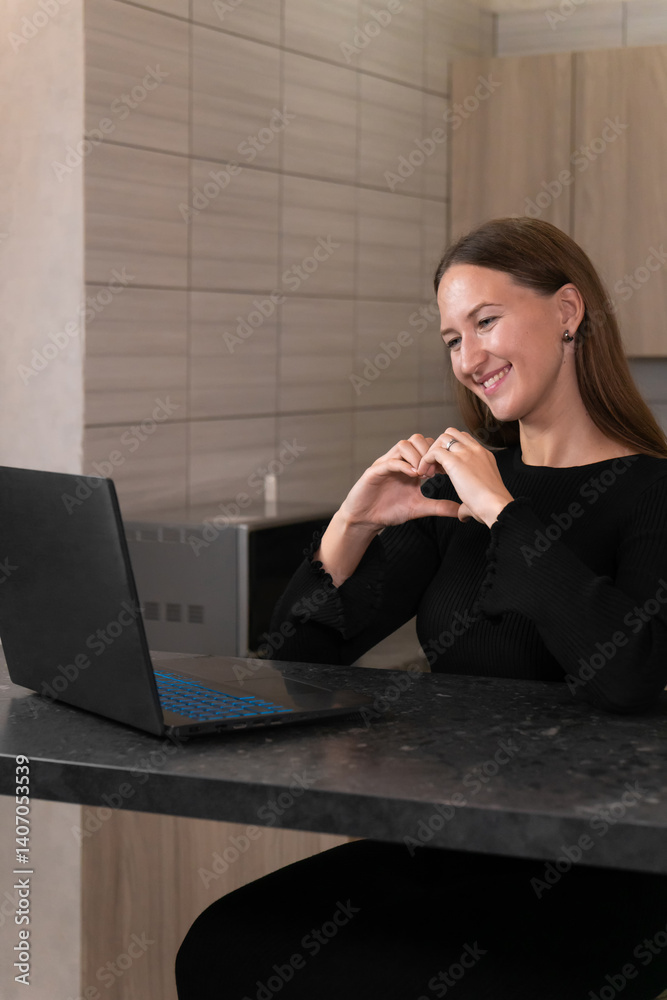 Smiling young woman is making a heart shape with her hands while video calling on her laptop, expressing love and affection to someone special. Vertical photo