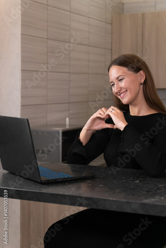 Obraz na płótnie Smiling young woman is making a heart shape with her hands while video calling on her laptop, expressing love and affection to someone special. Vertical photo w salonie