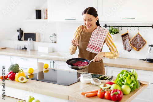 Canvastavla Pretty young lady frying meat and seasoning with salt, cooking dinner in kitchen