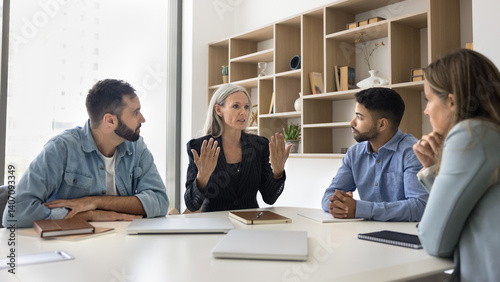 Mentorship and collaboration. Experienced senior female team leader boss business coach trainer speak talk to focused younger interns trainees diverse multicultural office workers at boardroom desk