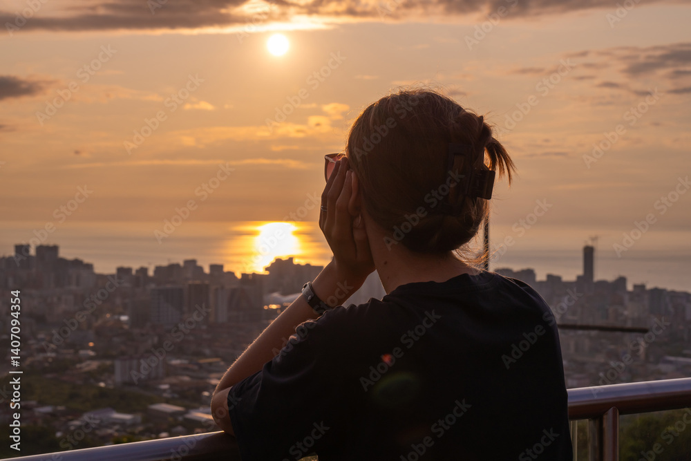 Young woman enjoys a peaceful view of the sunset over the city, leaning on the railing with her back to the camera, feeling calm and serene