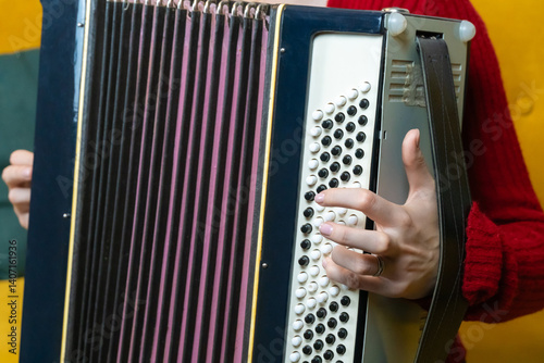 Woman musician in red sweater and dark pants playing a large black and white accordion while sitting on a yellow sofa in a bright room