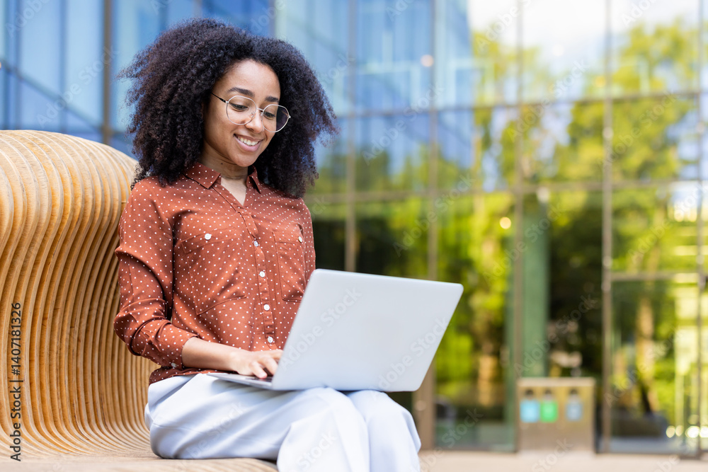A young woman with glasses smiles as she works on her laptop outside on a wooden bench in front of a modern building.