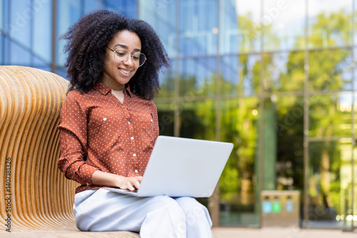 Obraz na płótnie A young woman with glasses smiles as she works on her laptop outside on a wooden bench in front of a modern building. w salonie