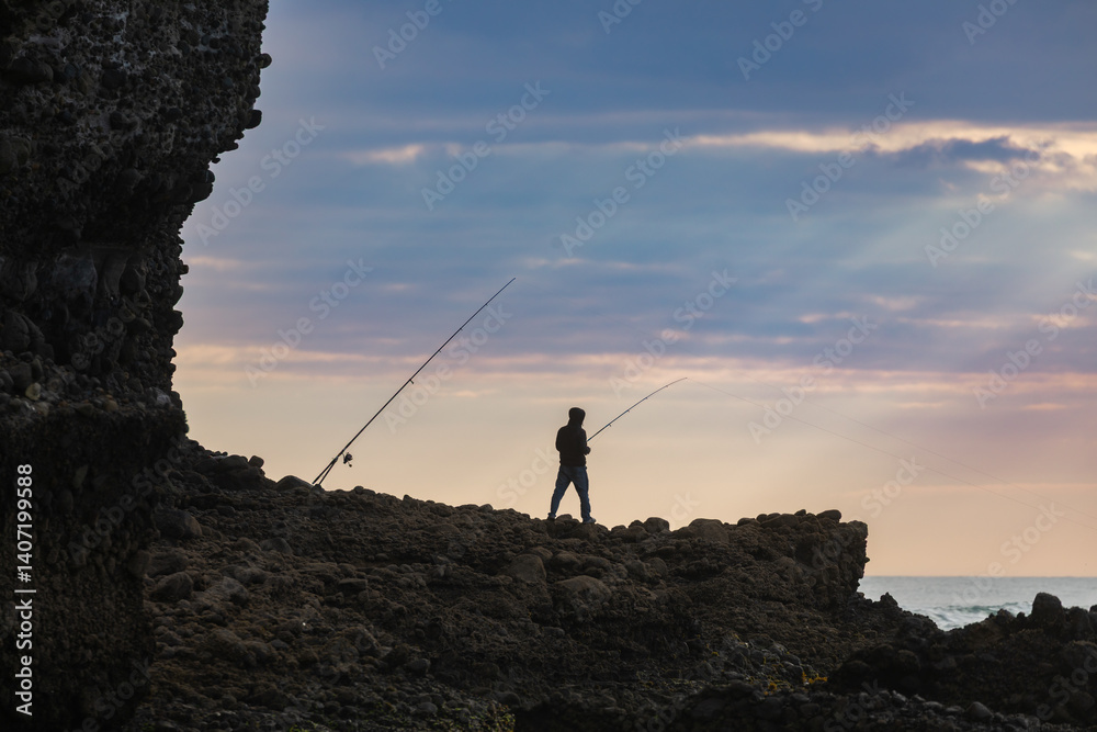 Obraz premium A lone fisherman casts his line from a rocky outcrop at dusk in Piha, Auckland, New Zealand. The silhouette of the man and his fishing rods stand out against the soft sky.