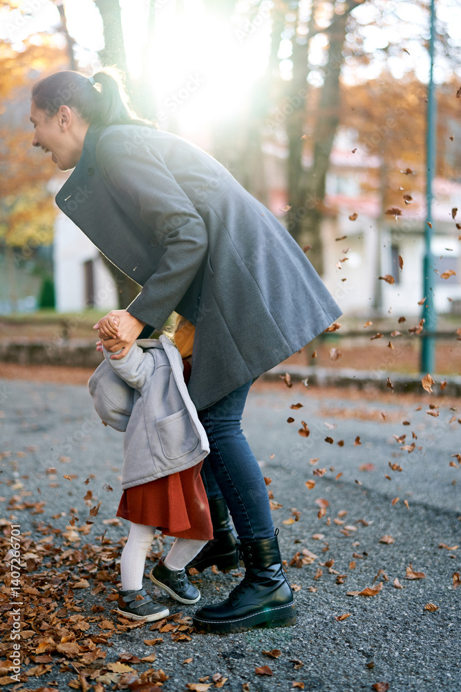 Smiling mom holding hands a little girl hiding her face in her lap while standing under falling leaves Smiling mom holding hands a little girl hiding her face in her lap while standing under falling leaves