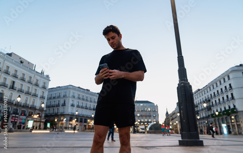 Obraz na płótnie Focused young man in black activewear checks fitness app on smartphone before urban jog at sunrise in city, surrounded by classic architecture and morning city energy w salonie