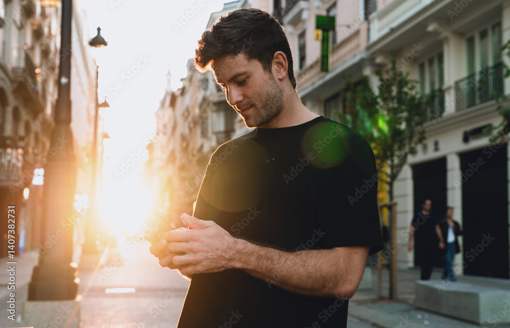 Young man in black shirt standing on sunlit street, focused on smartphone screen. Serene atmosphere blends modern lifestyle, morning routine and digital connectivity in city