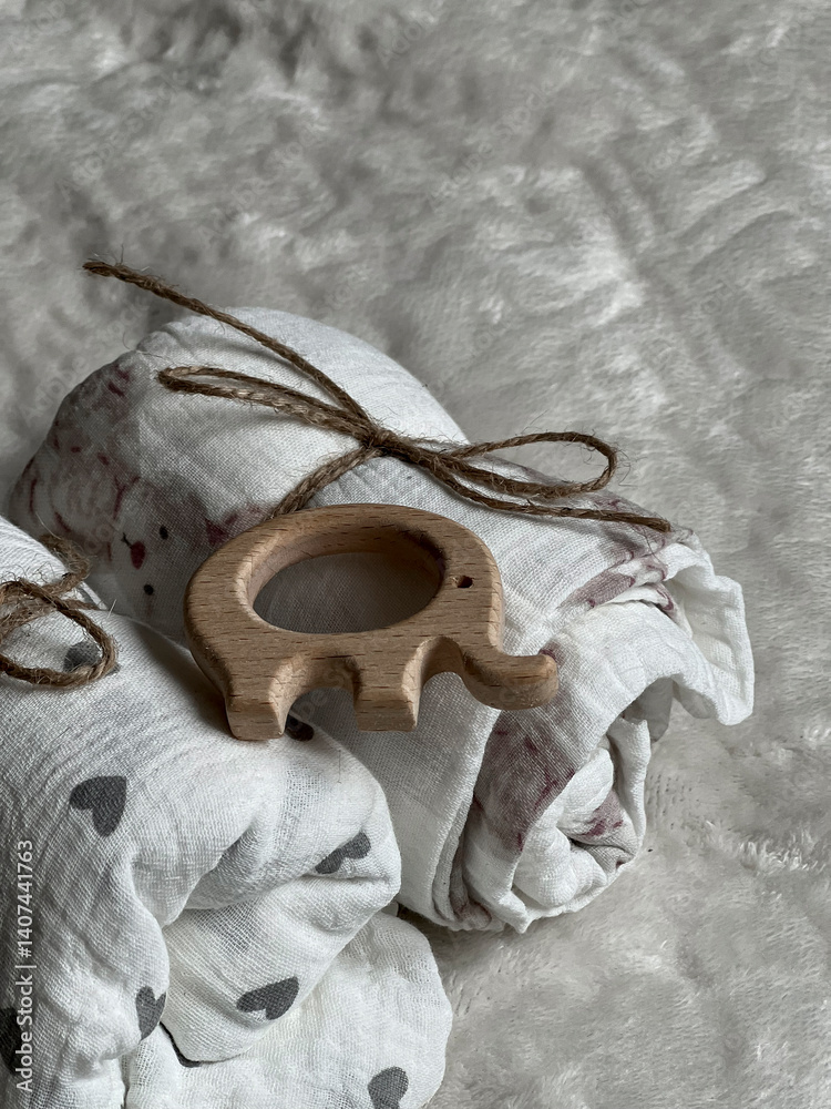 Two twisted muslin diapers and an elephant teether are lying on a milky-colored blanket. Children's fashion accessories