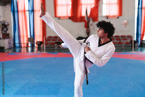 A male martial artist in a white uniform and black belt performs a high side kick on a blue mat inside a taekwondo gym with red curtains and chairs in the background