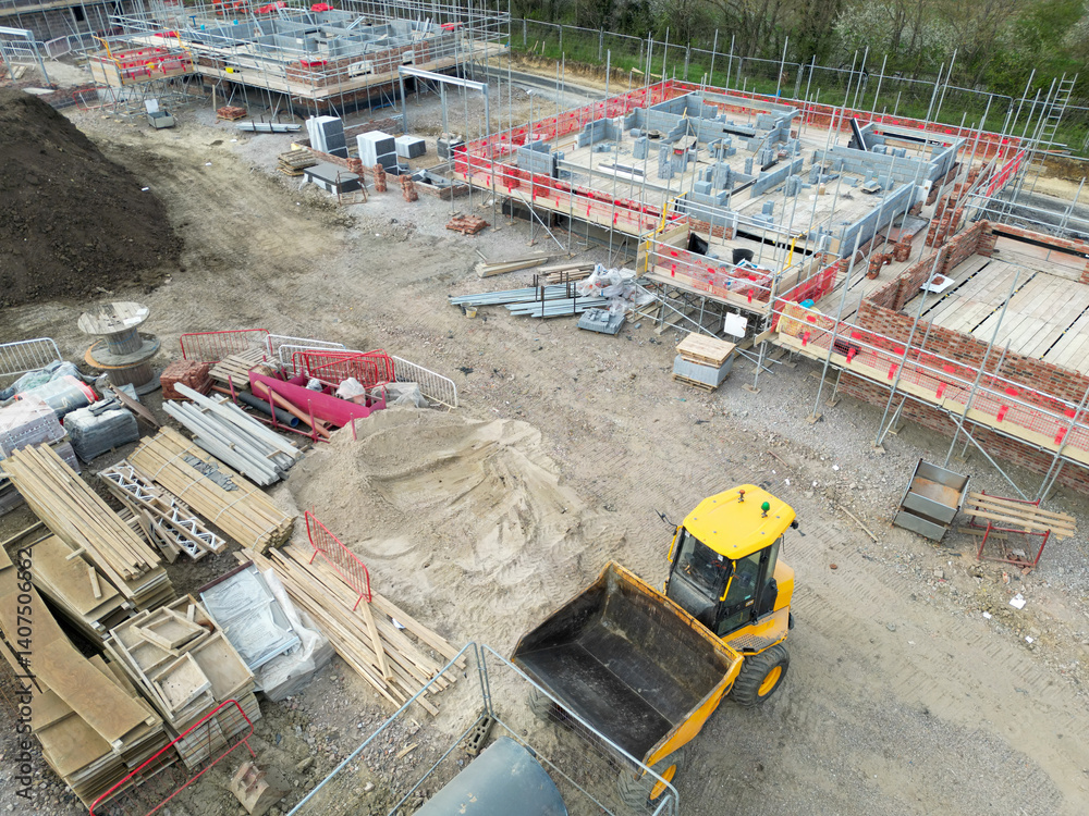 Aerial view of a parked up small digger and scoop located on a typical British housing development site. Partially built affordable and private homes are seen in the background.
