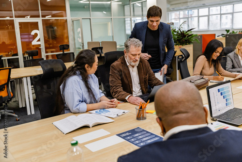 An overweight Asian woman in her 30s and a mid-30s White man, asking advice from their middle-aged White male colleague, as they're gathered around a table in an office hall, with other colleagues