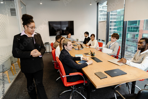 A Black female boss in her 30s, dressed in a black business suit, walking around the table in an office meeting room as her employees and colleagues all look at her, heading for her seat at the head