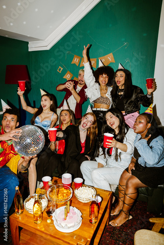 At a birthday party at home, many multiethnic stylish friends in their 20s cheerfully raise red cups while sitting and standing at a small table with snacks, one is a Latino man holding a disco ball.