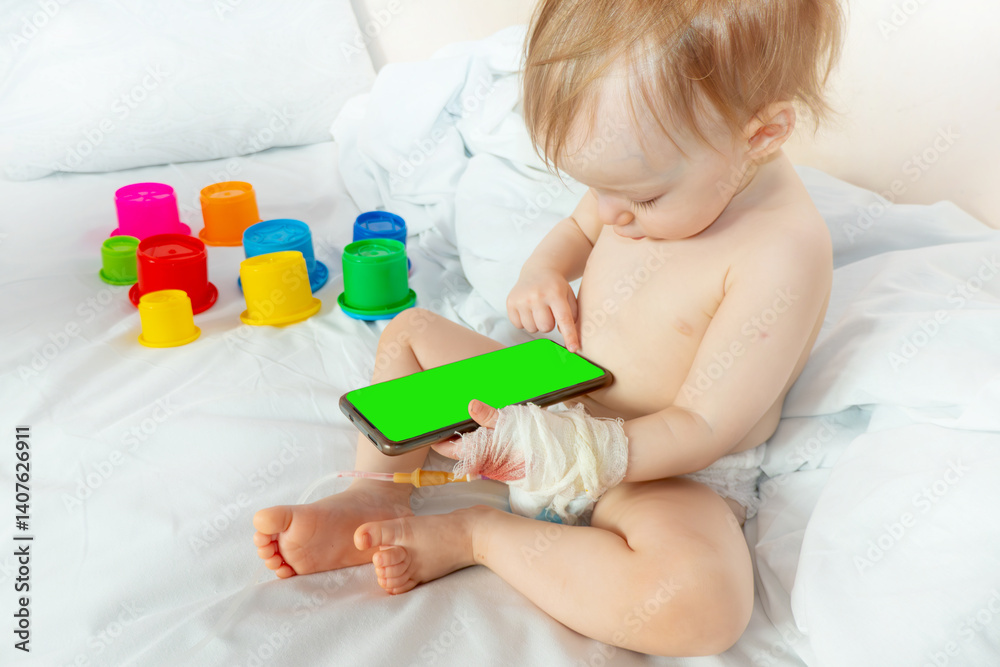 child in hospital sit in bed with mobile phone smiling. baby plays on the phone with intravenous infusion set to drop. concept of courage and joie de vivre. mockup with a green screen