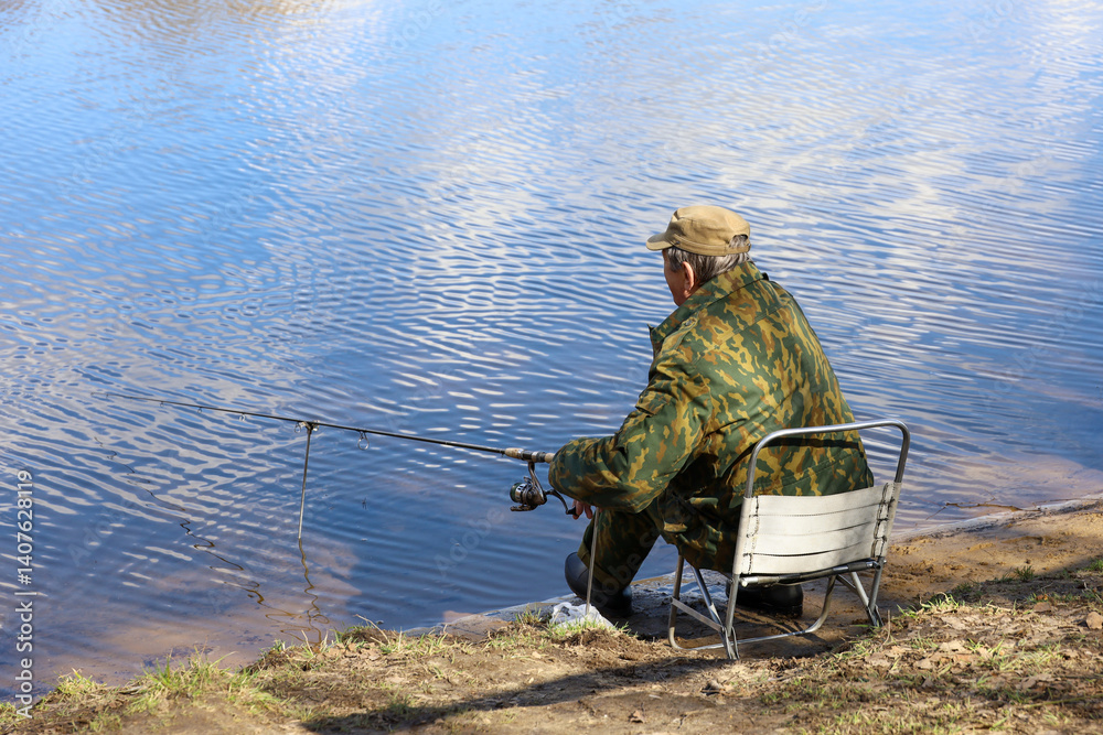 Fisherman sitting near the water with a fishing rod, side view. Man in camouflage angling on the lake coast at spring