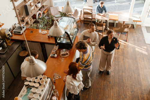An overhead view of a tall White man aged 20, with short, curly blond hair, placing an order in a small coffee shop as other customers and workers chat and work around him
