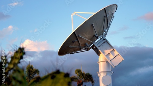 Large parabolic aerospace radar antenna pointing skyward, capturing signals from the cloudy atmosphere during a vibrant sunset, framed by palm trees in the foreground