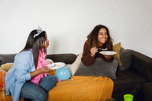 A Black woman aged 25, wearing a tiara on her head, sitting on a sofa at home and giggling together with her mid-20s Latina female friend, as they're eating cake and celebrating her birthday photo