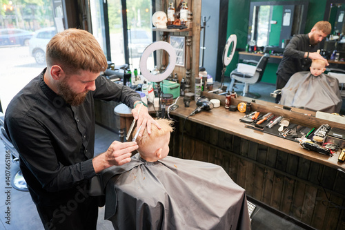 Man barber cutting little boy's hair using comb and scissors. Child getting haircut from adult male, likely barber. Professional hairdresser and cute client at modern barbershop.