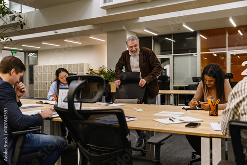 A team collaborates in a modern office, with a senior colleague smiling and fostering a positive atmosphere as he takes a seat. A concept of teamwork, joy, and leadership.