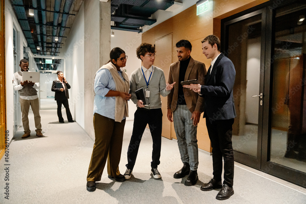 Fototapeta premium A diverse group of adult men and women, including Indian, Black, White, and Middle Eastern coworkers, discuss in a modern office hallway with tablets and notebooks.