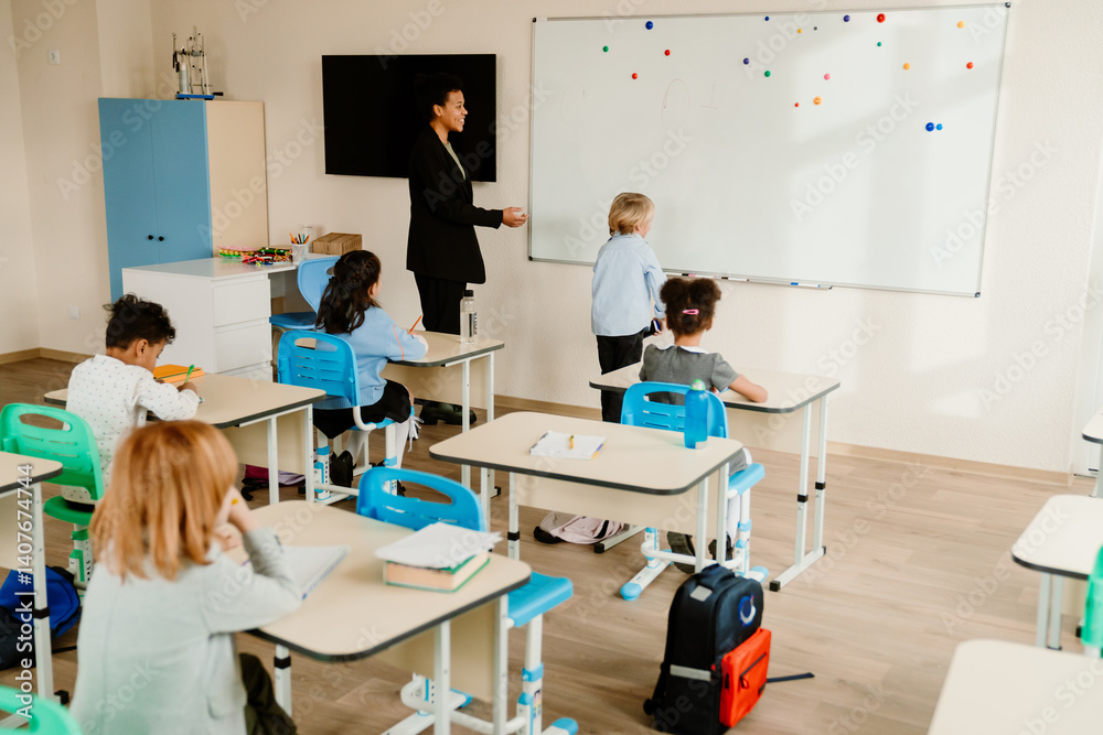 Obraz premium During a lesson in a classroom, a young Black teacher and a White boy stand at the whiteboard, while other children in the class sit at their desks. Concept of school activities, a study process.