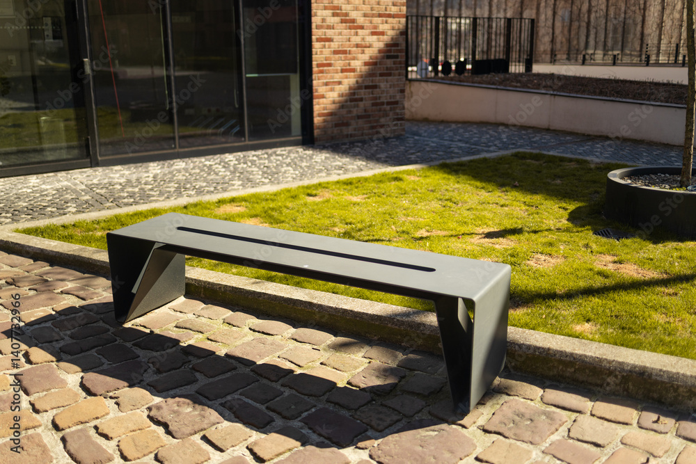 A sleek, modern bench placed in a green urban park with a clean design. The bench is surrounded by cobblestones and grass, providing a contemporary setting in the city’s outdoor space.