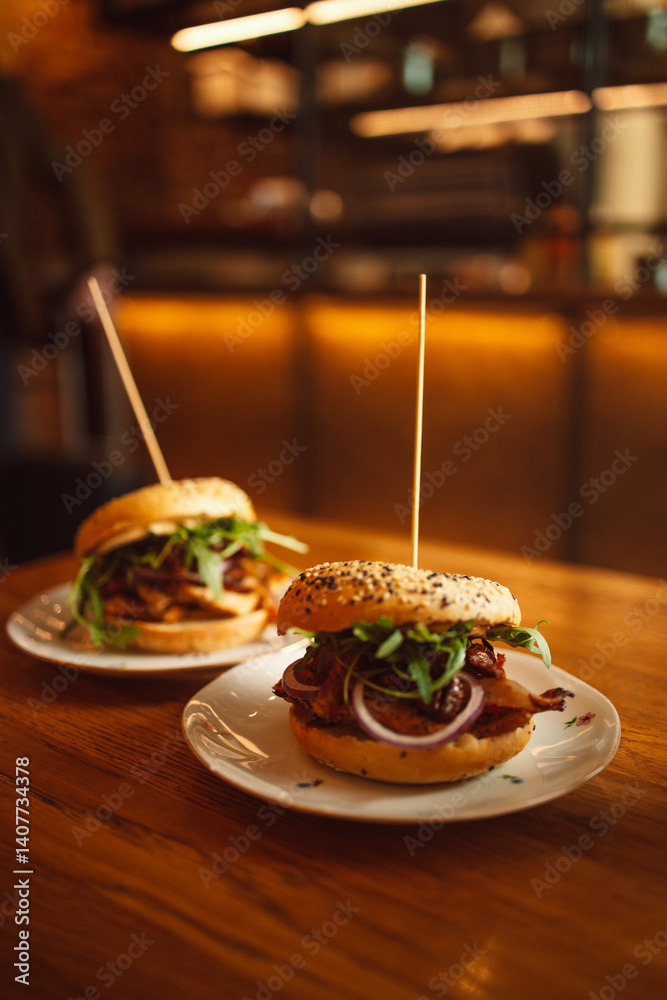 Two delicious gourmet burgers on white plates, topped with crispy bacon, fresh greens, and onions, placed on a wooden table. The soft sesame buns are skewered with a toothpick for easy handling. The