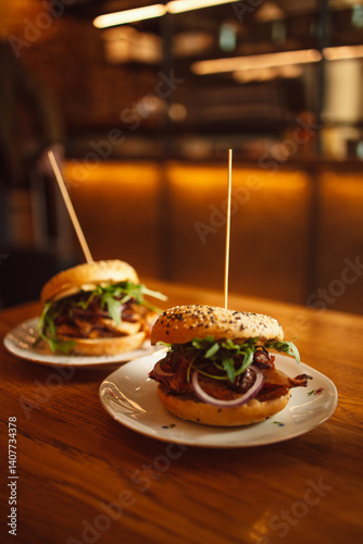 Obraz na płótnie Two delicious gourmet burgers on white plates, topped with crispy bacon, fresh greens, and onions, placed on a wooden table. The soft sesame buns are skewered with a toothpick for easy handling. The w salonie