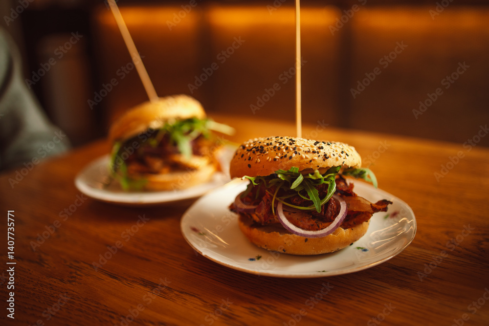 Two delicious gourmet burgers on white plates, topped with crispy bacon, fresh greens, and onions, placed on a wooden table. The soft sesame buns are skewered with a toothpick for easy handling. The
