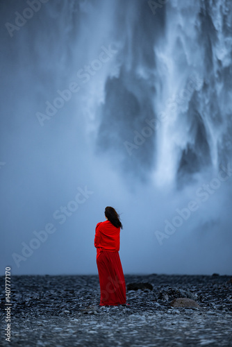 A woman in a red dress stands before the powerful Skógafoss waterfall in Iceland, her figure contrasting sharply against the mist and roaring cascade.