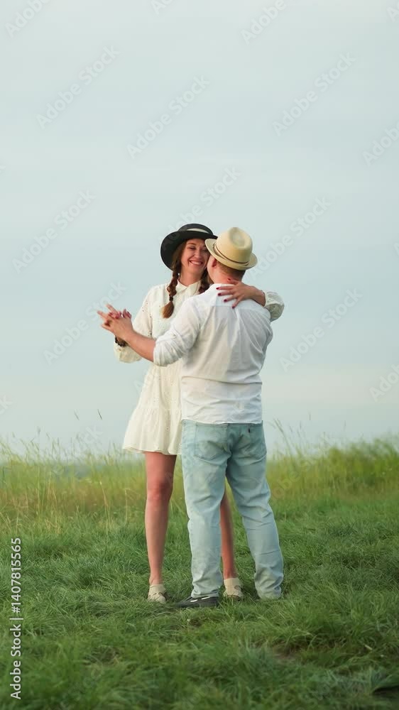 A loving couple dances together on a grassy hill, with the man wearing a white shirt, hat, and jeans, and the woman dressed in a white dress and black hat.The scene captures a tender and joyful moment A loving couple dances together on a grassy hill, with the man wearing a white shirt, hat, and jeans, and the woman dressed in a white dress and black hat.The scene captures a tender and joyful moment