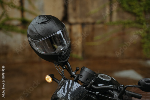 A sleek black motorcycle helmet with rain drops, emphasizing protection and style, sits on a bike in an urban setting under a rustic backdrop.