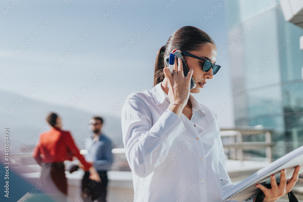 Businesswoman in sunglasses talking on phone and holding documents on a high rise balcony. Two other business people are seen in the background, with a cityscape view.