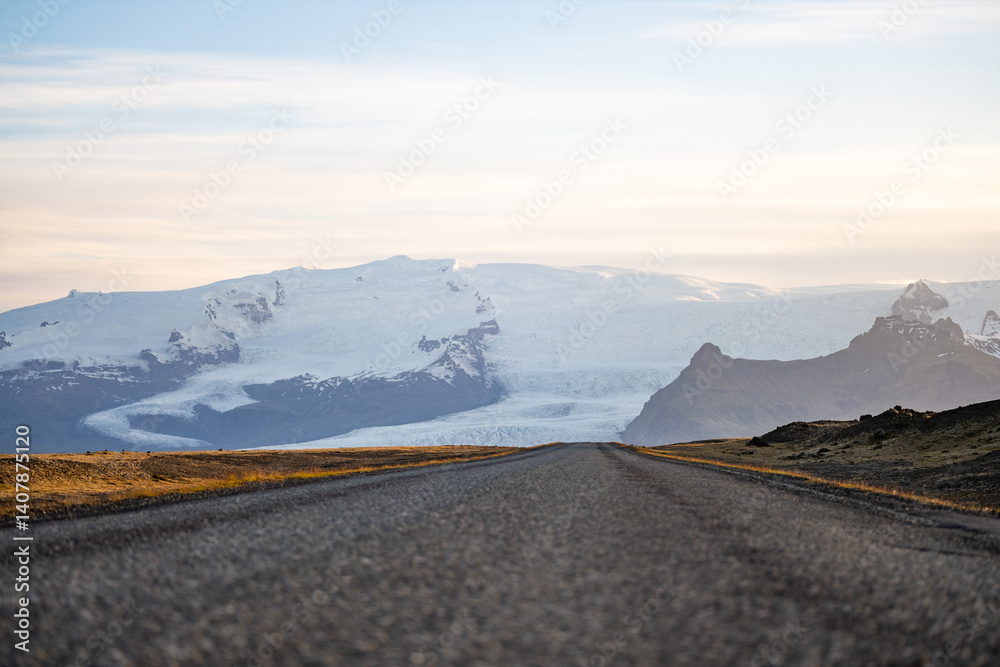 A long empty road leads toward a majestic glacier-covered mountain in Iceland, under a soft pastel sky at sunrise. A breathtaking view of nature's raw beauty and endless adventure.