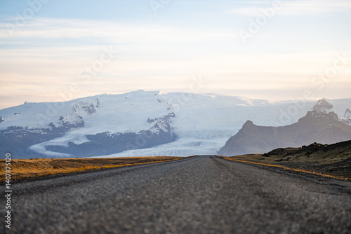 A long empty road leads toward a majestic glacier-covered mountain in Iceland, under a soft pastel sky at sunrise. A breathtaking view of nature's raw beauty and endless adventure.