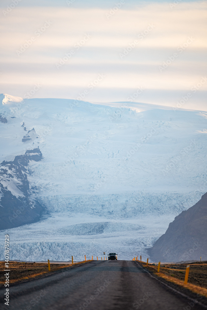 A long empty road leads toward a majestic glacier-covered mountain in Iceland, under a soft pastel sky at sunrise. A breathtaking view of nature's raw beauty and endless adventure.