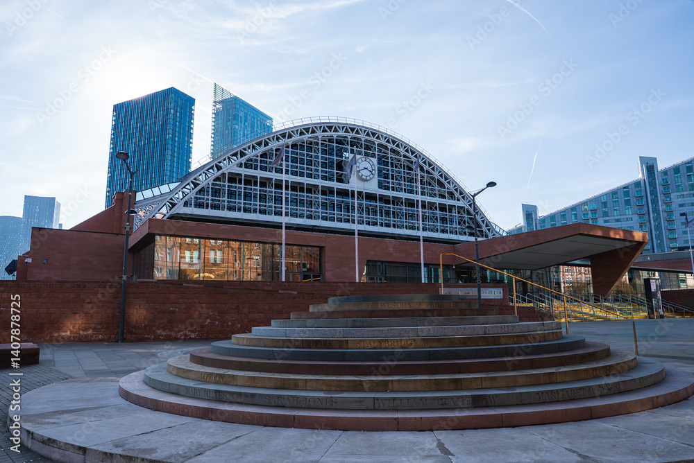 View of the Manchester Central Convention Complex with its arched roof and clock, surrounded by modern skyscrapers and a tiered stone structure.