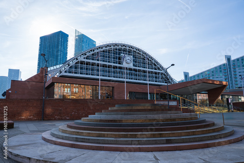 View of the Manchester Central Convention Complex with its arched roof and clock, surrounded by modern skyscrapers and a tiered stone structure.