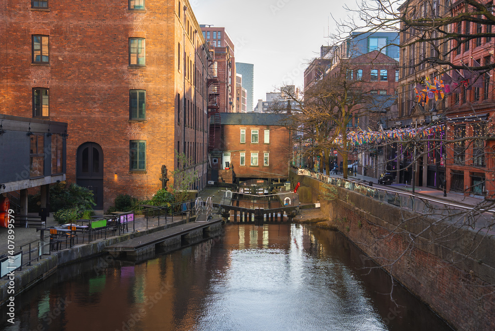 A canal in Manchester, England, with a central lock, historic red brick buildings, modern structures, colorful flags, and outdoor seating areas.