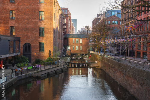 Obraz na płótnie A canal in Manchester, England, with a central lock, historic red brick buildings, modern structures, colorful flags, and outdoor seating areas. w salonie