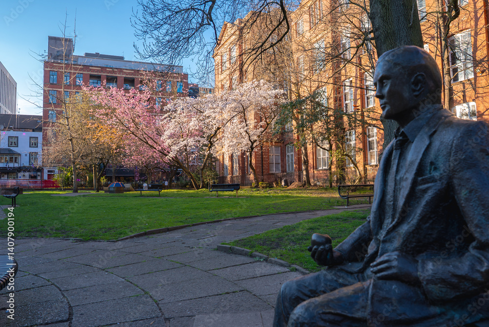 A serene park in Manchester featuring a bronze statue, blooming cherry blossoms, benches, pathways, and red brick and modern buildings in the background.