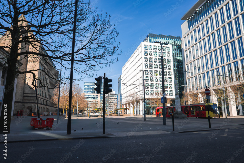 A Manchester street scene with a stone building on the left, glass structures on the right, a red bus, bare trees, a traffic light, and a clear blue sky.