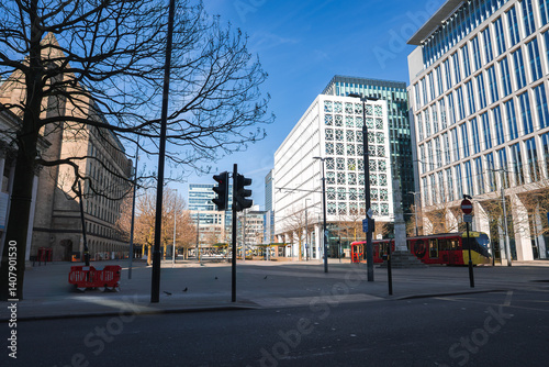 Obraz na płótnie A Manchester street scene with a stone building on the left, glass structures on the right, a red bus, bare trees, a traffic light, and a clear blue sky. w salonie