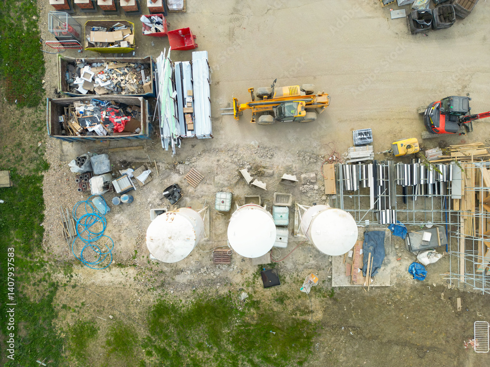 Drone top down view of a housing construction site. A single yellow tele hoist vehicle is parked ip near three tall white mortar silos at the lower part of the image.