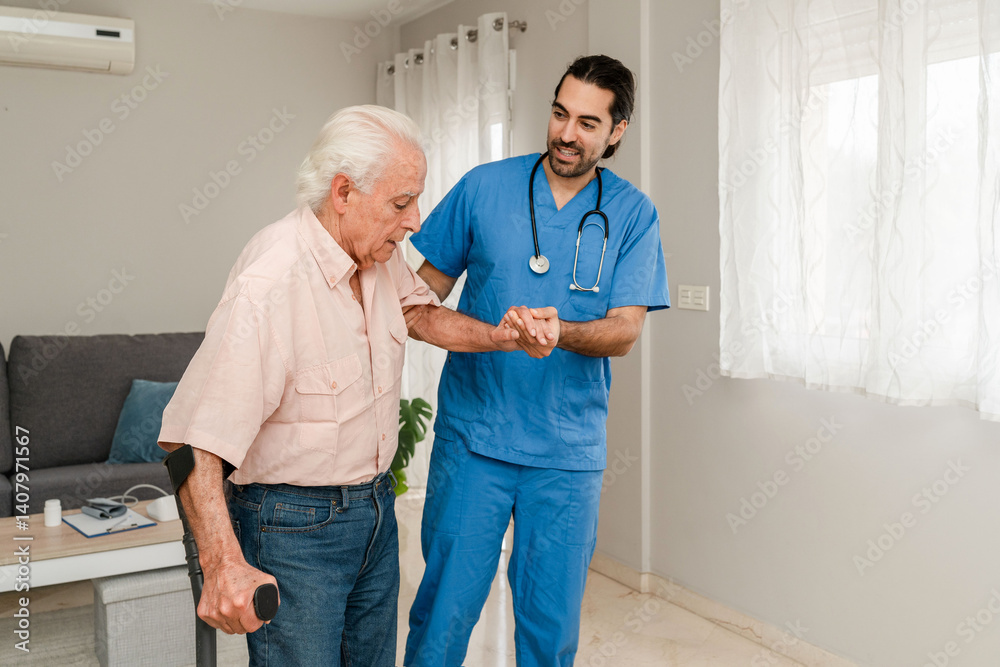 Fototapeta premium Kind male nurse in blue uniform helping a senior man with crutches during home visit