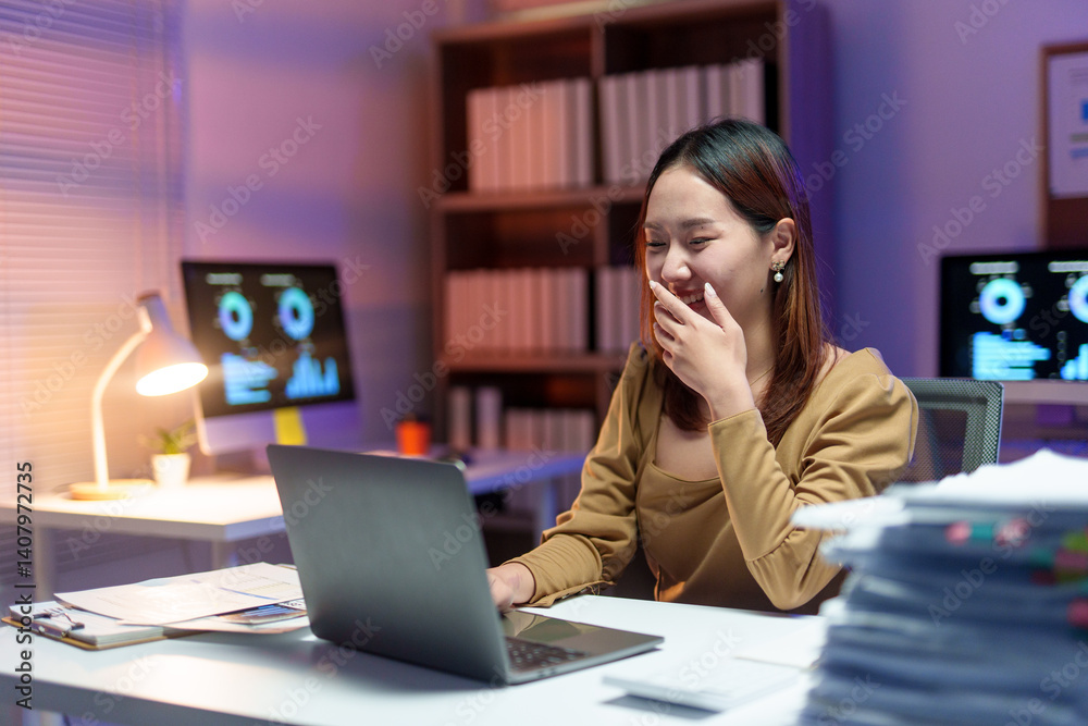 Young happy businesswoman laughing while using laptop, working late at night in office with computer and paperwork Young happy businesswoman laughing while using laptop, working late at night in office with computer and paperwork