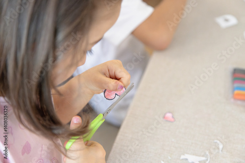 During a creative activity at home, a young child carefully uses safety scissors to cut out a heart shape, developing fine motor skills, autonomy, and concentration.